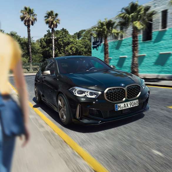 BMW M135i xDrive, vista frontal de tres cuartos. BMW M135i xDrive en Black Sapphire metallic; vista frontal de tres cuartos en movimiento, con una mujer en primer plano.