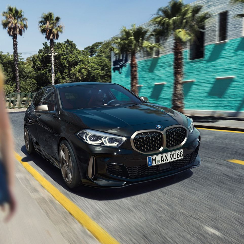 BMW M135i xDrive, vista frontal de tres cuartos. BMW M135i xDrive en Black Sapphire metallic; vista frontal de tres cuartos en movimiento, con una mujer en primer plano.