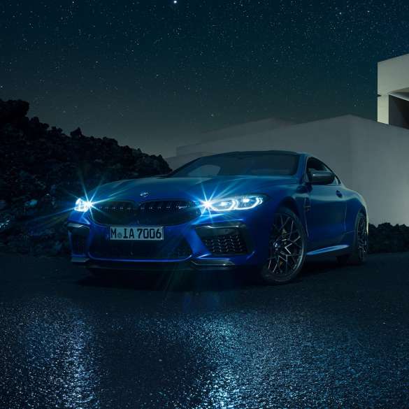 BMW M8 Competition Coupé, vista frontal tres cuartos. BMW M8 Competition Coupé en Marina Bay Blue metálico con luces LED brillantes, vista frontal tres cuartos en alto frente a una villa de noche.