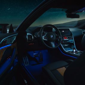 Interior BMW M8 Competition Coupé. BMW M8 Competition Coupé, interior de noche con volante en cuero M y luz ambiente azul con la puerta de conductor abierta.