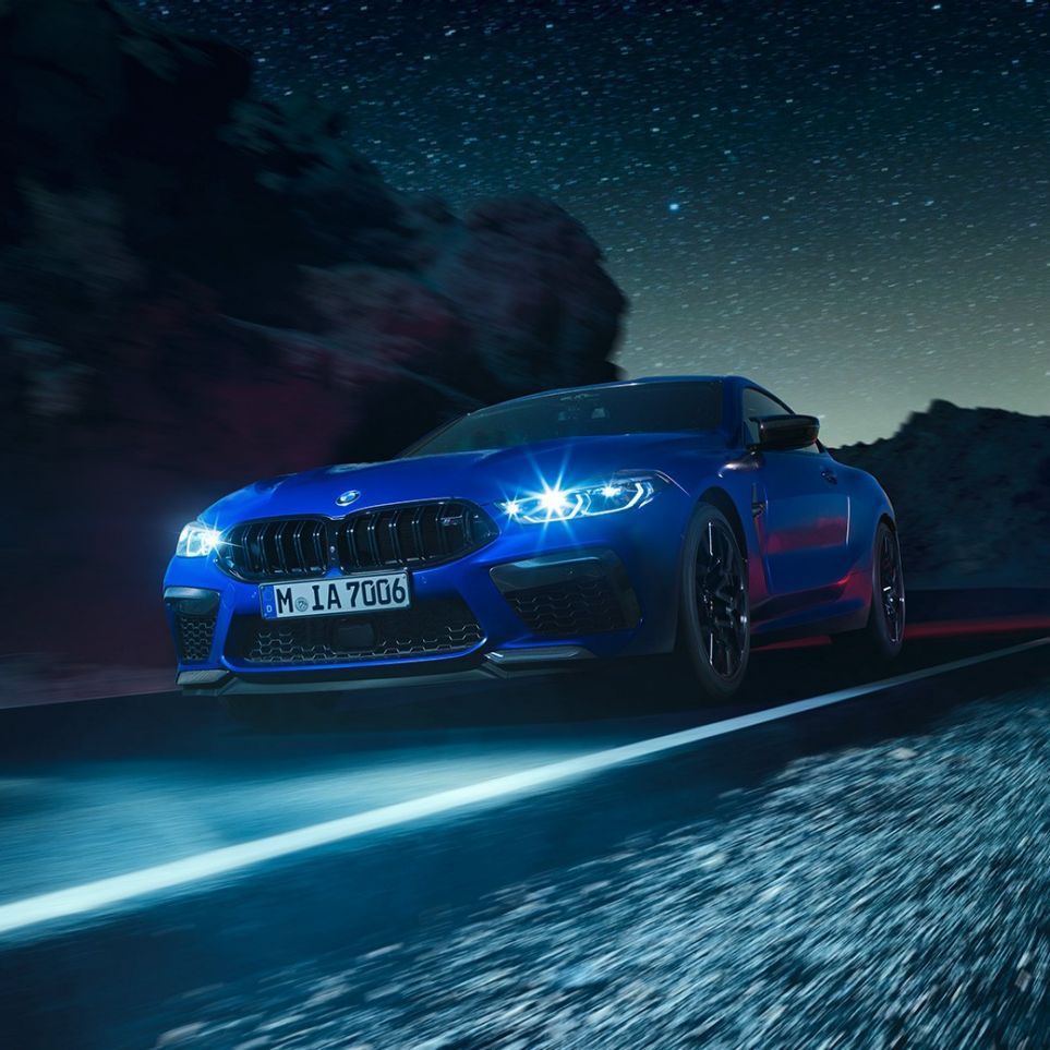 Conduciendo el BMW M8 Competition Coupé, vista frontal tres cuartos. BMW M8 Competition Coupé en Marina Bay Blue metálico con luces LED iluminadas, vista frontal tres cuartos, conduciendo de noche.