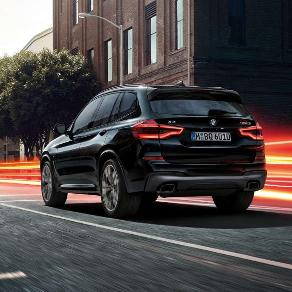 BMW X3 M40i, vista trasera de tres cuartos. BMW X3 M40i en Black Sapphire metálico, en movimiento, exterior, vista trasera de tres cuartos frente a un fondo urbano con franjas de luz.