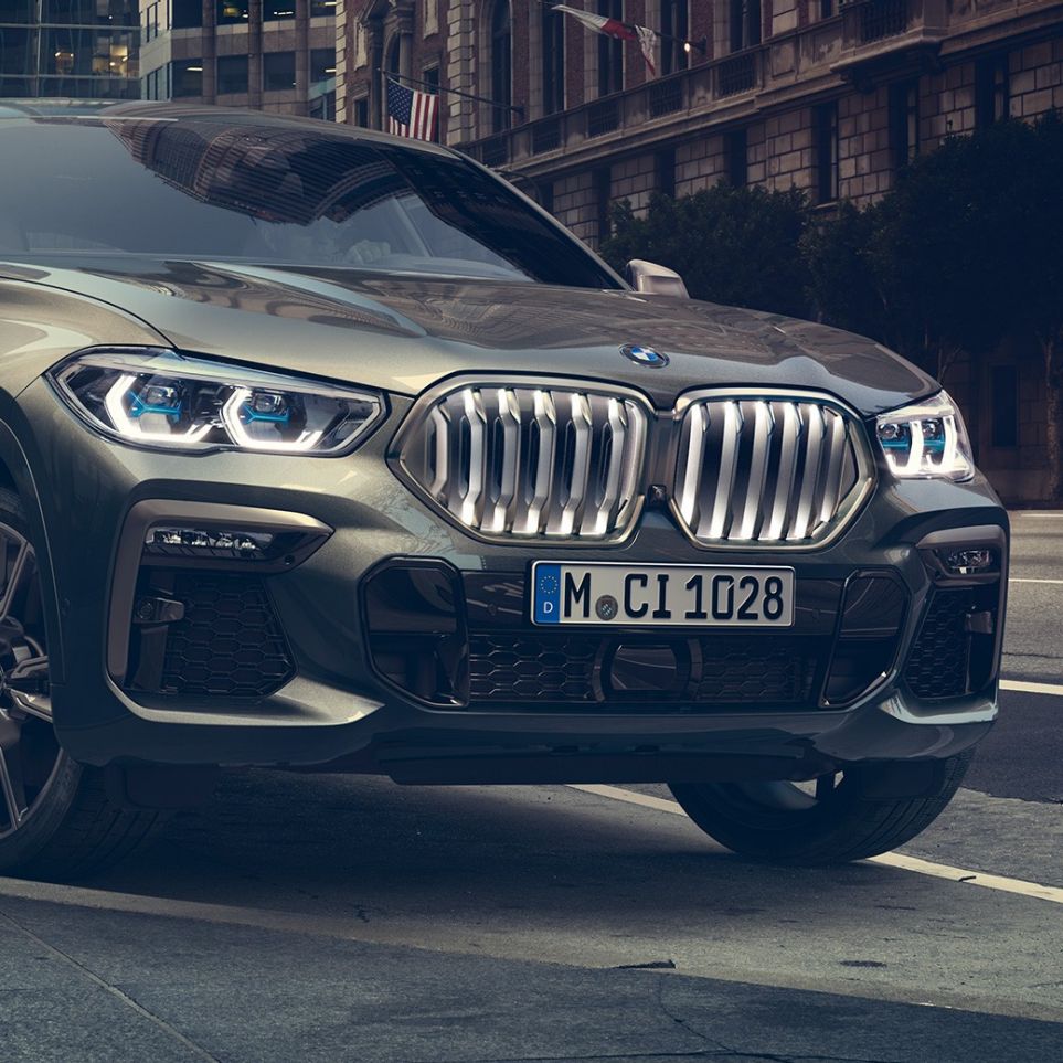 BMW X6 M50i parado BMW X6 M50i en perspectiva frontal de tres cuartos parado frente a un escenario urbano.