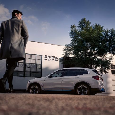 BMW iX3 G08 Vista lateral delante de edificio con un hombre BMW iX3 G08 2020 Mineralweiss metalizado Vista lateral delante de un edificio con un hombre