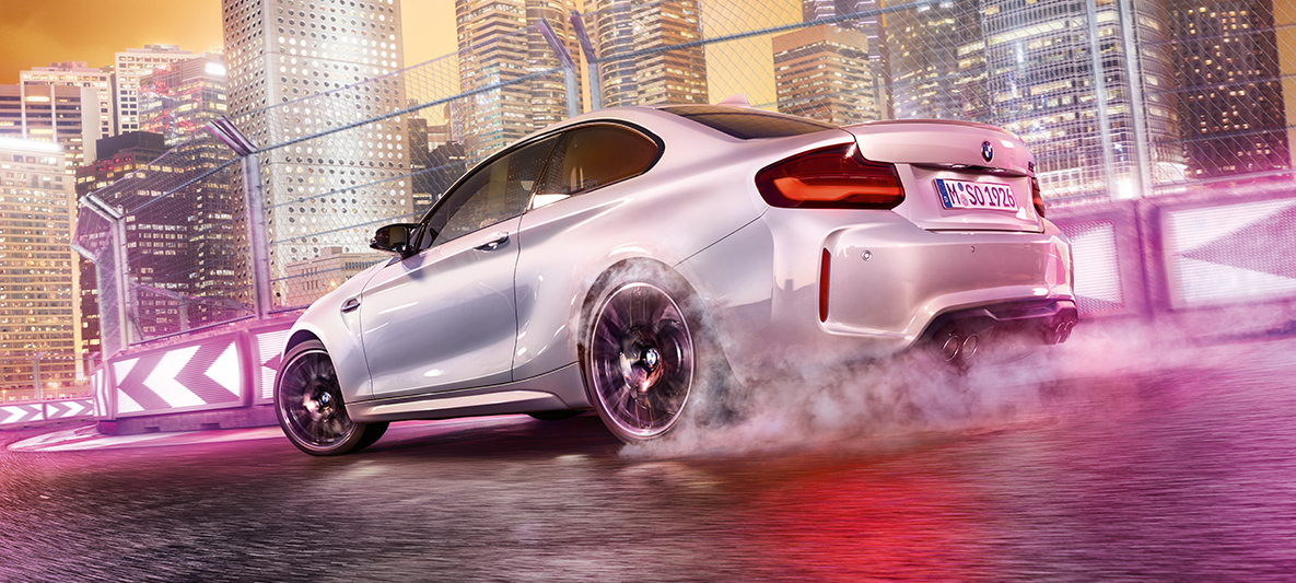 BMW M2 Competition acelera BMW M2 Competition blanco en la pista de carrera frente al paisaje nocturno de la ciudad.