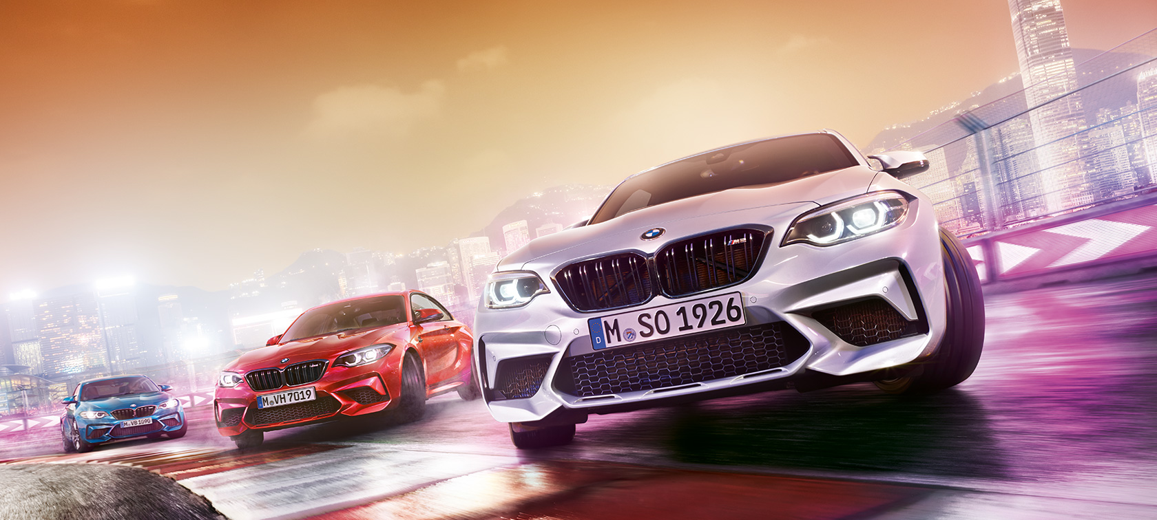 BMW M2 Competition en acción Tres BMW M2 Competition en la pista de carrera frente al paisaje nocturno de la ciudad.
