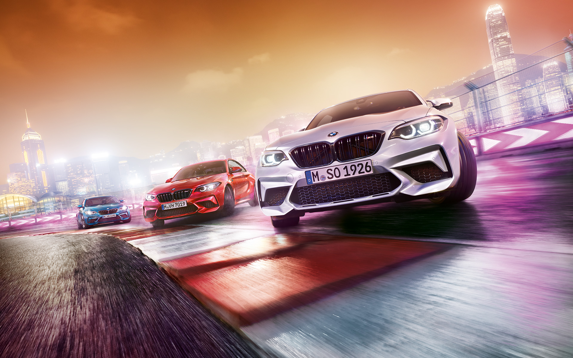 BMW M2 Competition en acción Tres BMW M2 Competition en la pista de carrera frente al paisaje nocturno de la ciudad.