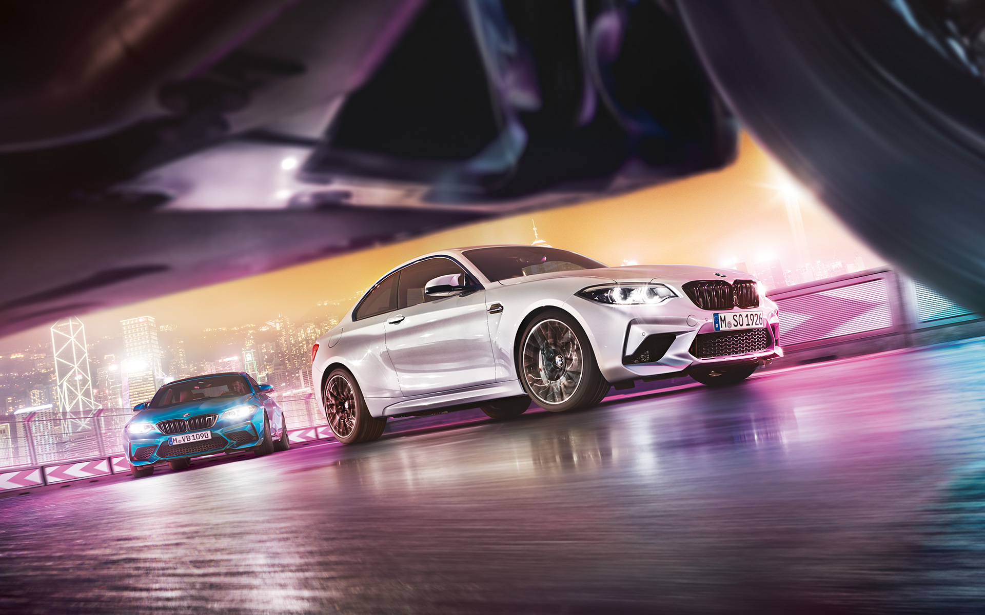 BMW M2 Competition en el asfalto. Dos BMW M2 Competition en la pista de carrera frente al paisaje nocturno de la ciudad a nivel de calle.