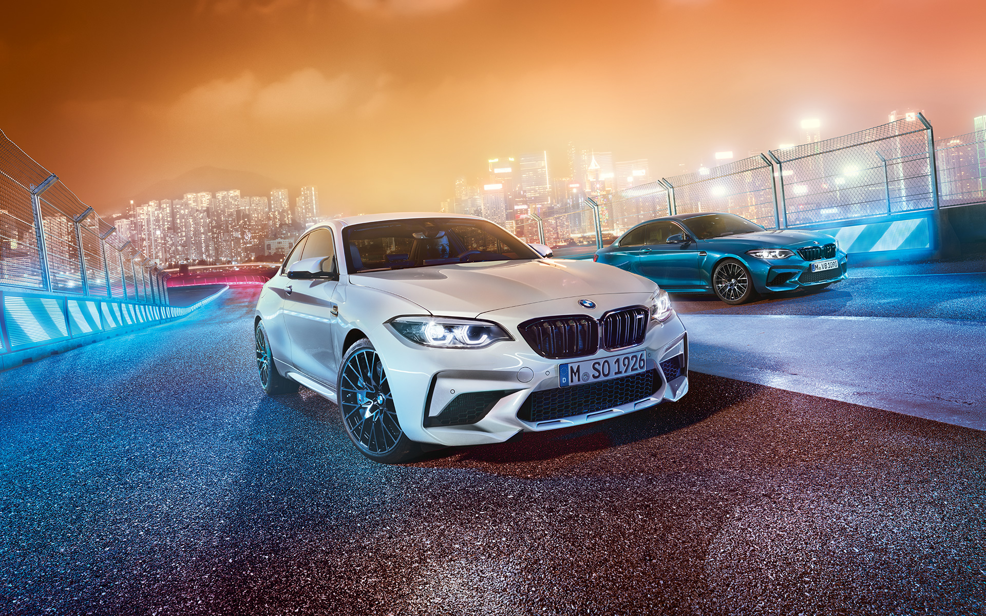 Dos BMW M2 Competition en la pista de carrera. BMW M2 Competition, blanco y negro, en la pista de carrera frente al paisaje nocturno de la ciudad.