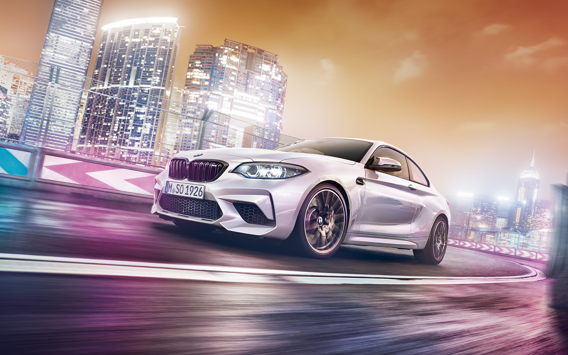 BMW M2 Competition blanco, con paisaje. BMW M2 Competition blanco en la pista de carrera frente al paisaje nocturno de la ciudad.