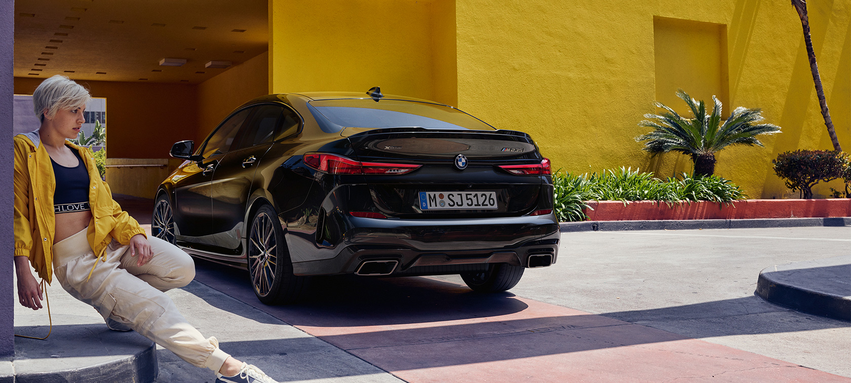 Vista trasera de tres cuartos del BMW M235i xDrive Gran Coupé Vista trasera de tres cuartos del BMW M235i xDrive Gran Coupé Black Sapphire metálico F44 con una mujer al frente