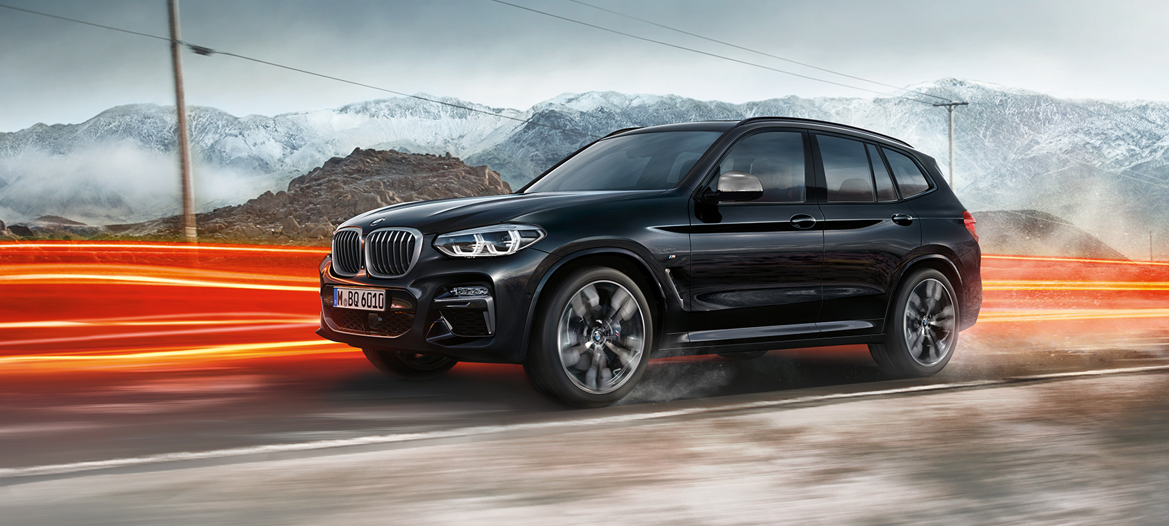 BMW X3 M40i, vista lateral. BMW X3 M40i en Black Sapphire metálico, en movimiento, exterior, vista lateral frente a un paisaje montañoso con franjas de luz.