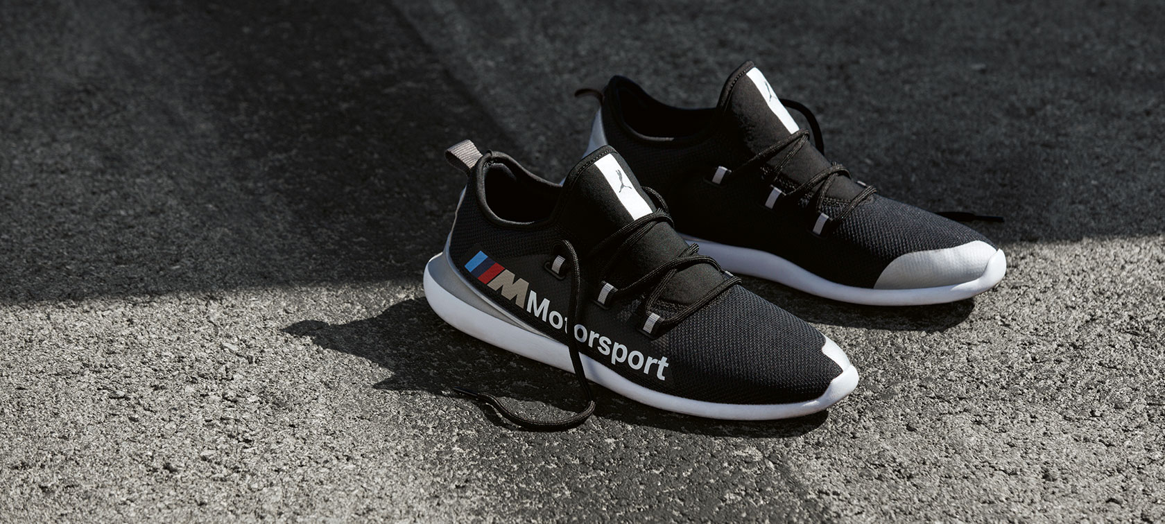 Tenis PUMA EVO CAT BMW M Motorsport. Un par de tenis PUMA EVO CAT BMW M Motorsport de la colección BMW M Motorsport se muestra de pie sobre una superficie de asfalto.
