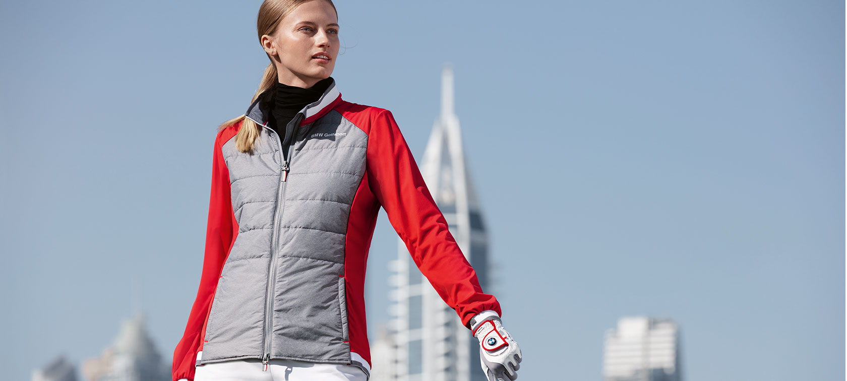 Chamarra BMW Golfsport para mujer. La joven de la foto presenta artículos de primera calidad de la colección BMW Golfsport con el telón de fondo del horizonte de una gran ciudad.
