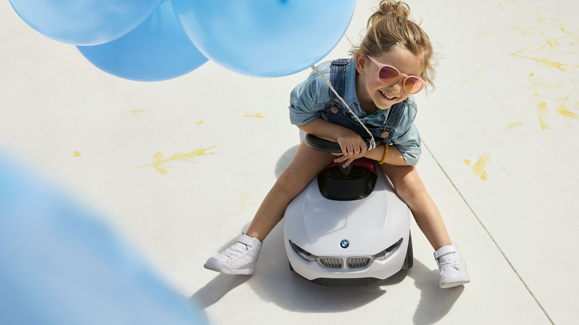 Colección BMW Kids. Una niña con lentes de sol rosas y un globo azul se sienta feliz en un BMW Baby Racer.