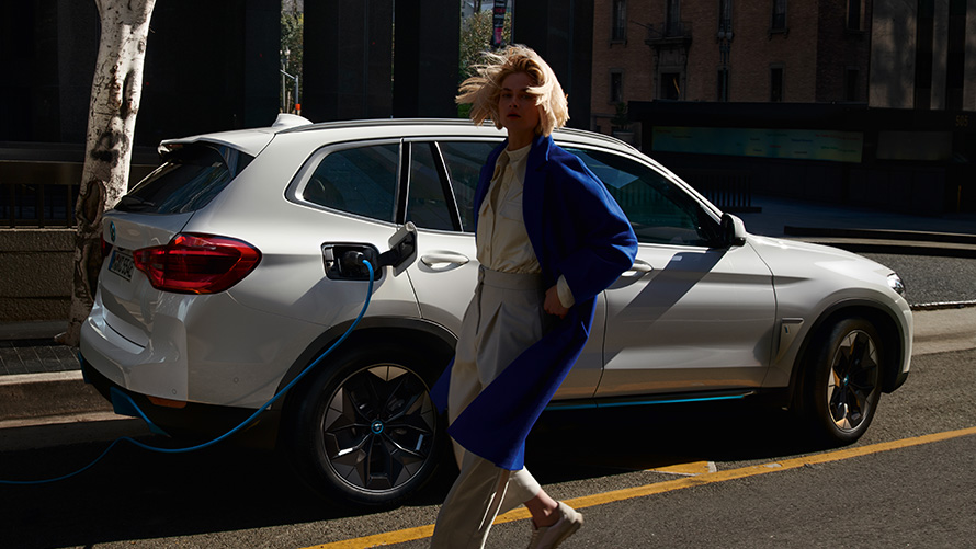 Mujer delante de un BMW iX3 conectado a un punto de carga BMW Financial Services Vehículo eléctrico BMW Híbrido Leasing Mujer delante de un BMW iX3 conectado a un punto de carga