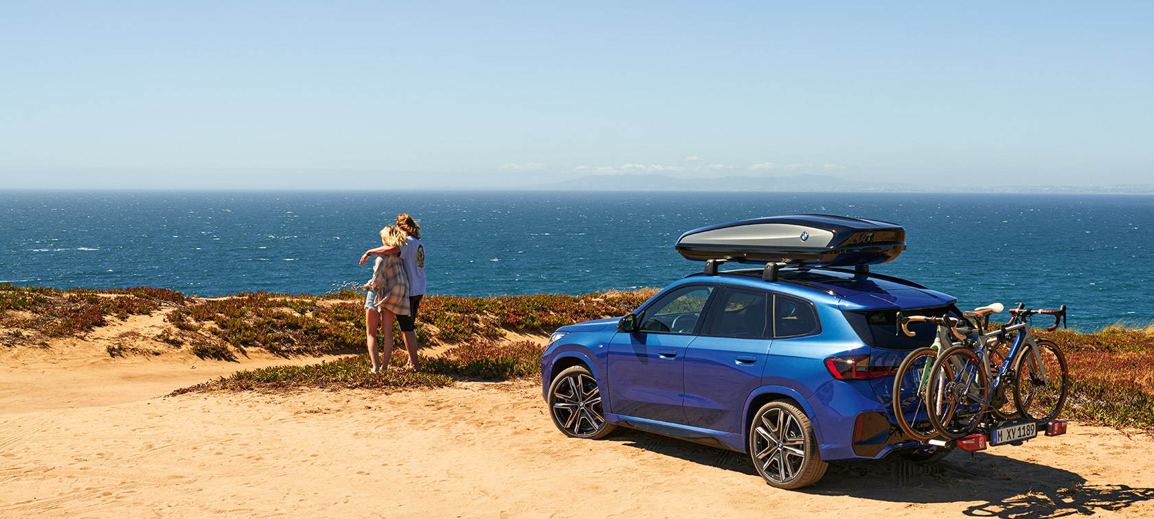 Accesorios Originales BMW para primavera y verano BMW X1 U11 parado sobre la arena frente a la costa con 2 personas